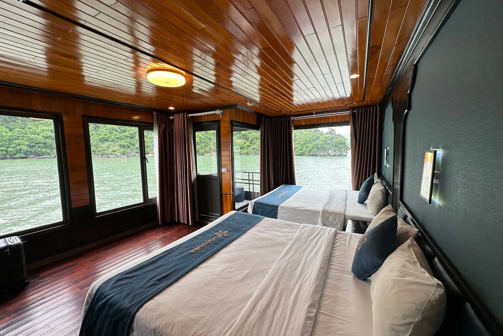 A wooden cabin interior with two beds, large windows showing water and karst islands outside, and text embroidered on the bed runners.