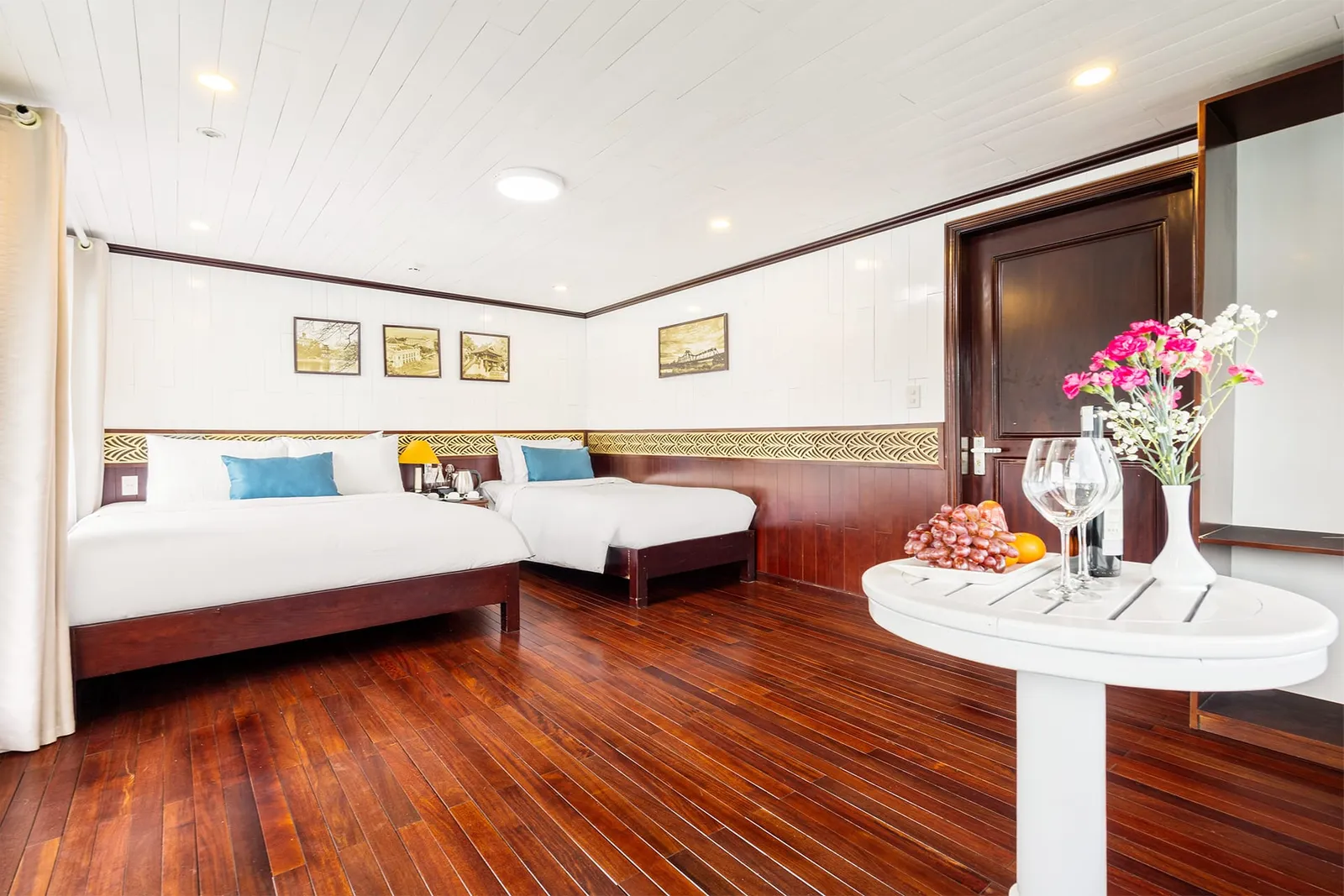A cabin with two double beds, dark red wooden floor, white paneled walls, framed pictures, and a round white table holding fruit, wine glasses, a bottle and pink flowers.