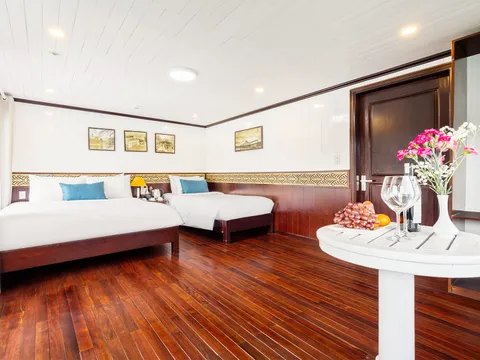 A cabin with two double beds, dark red wooden floor, white paneled walls, framed pictures, and a round white table holding fruit, wine glasses, a bottle and pink flowers.