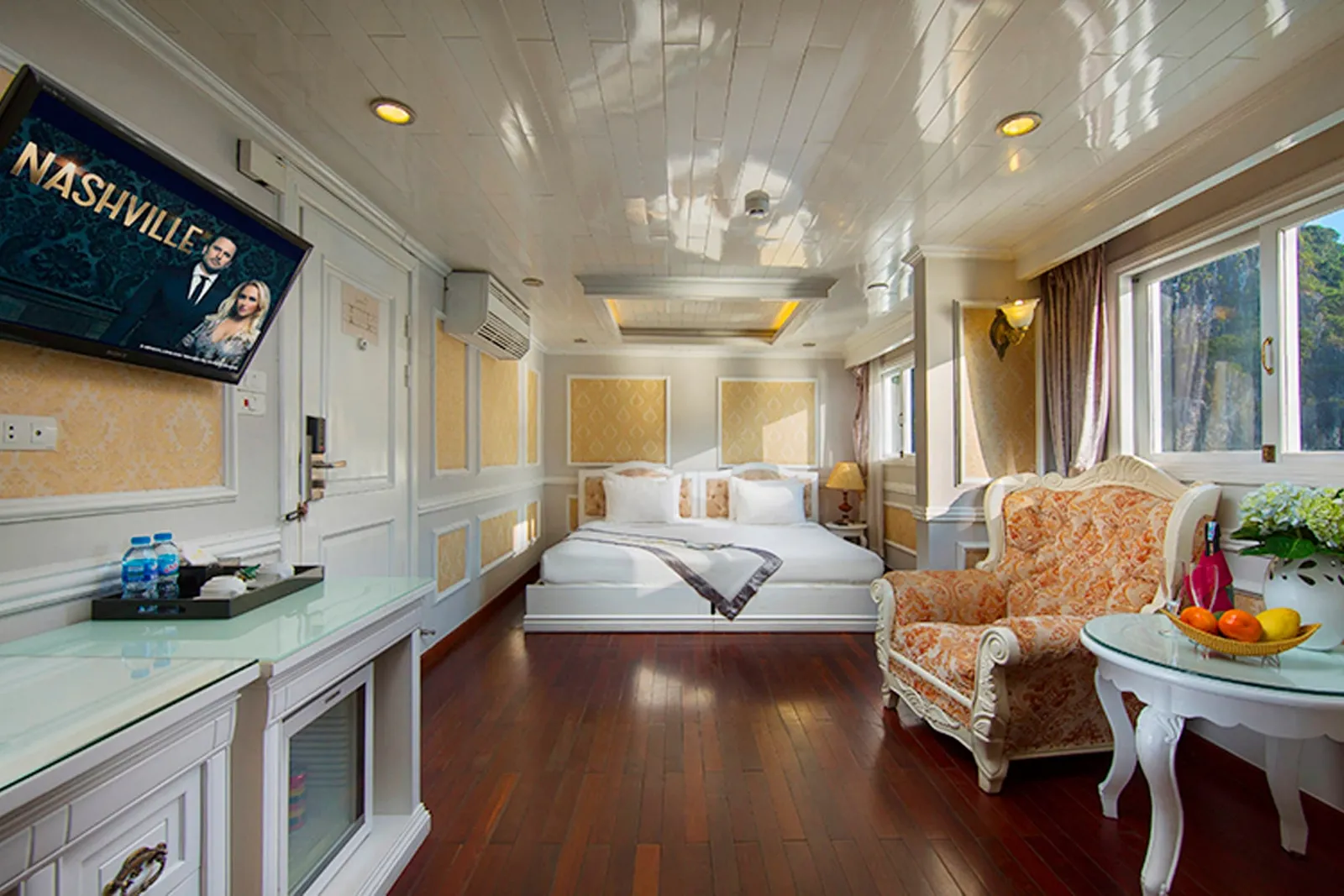 A cabin with a double bed, ornate armchair, fruit bowl on a glass table, wall-mounted TV showing a Nashville show, and windows looking out onto green limestone karst cliffs.