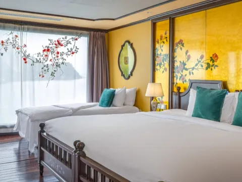 A cabin with a large bed with teal cushions, a twin bed, gold walls painted with floral branches, a TV and desk, and a window looking out onto a hazy karst-like landform.