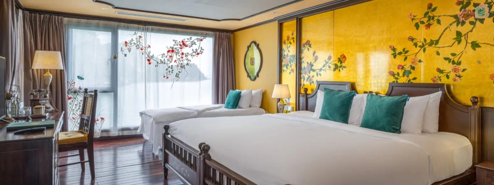 A cabin with a large bed with teal cushions, a twin bed, gold walls painted with floral branches, a TV and desk, and a window looking out onto a hazy karst-like landform.