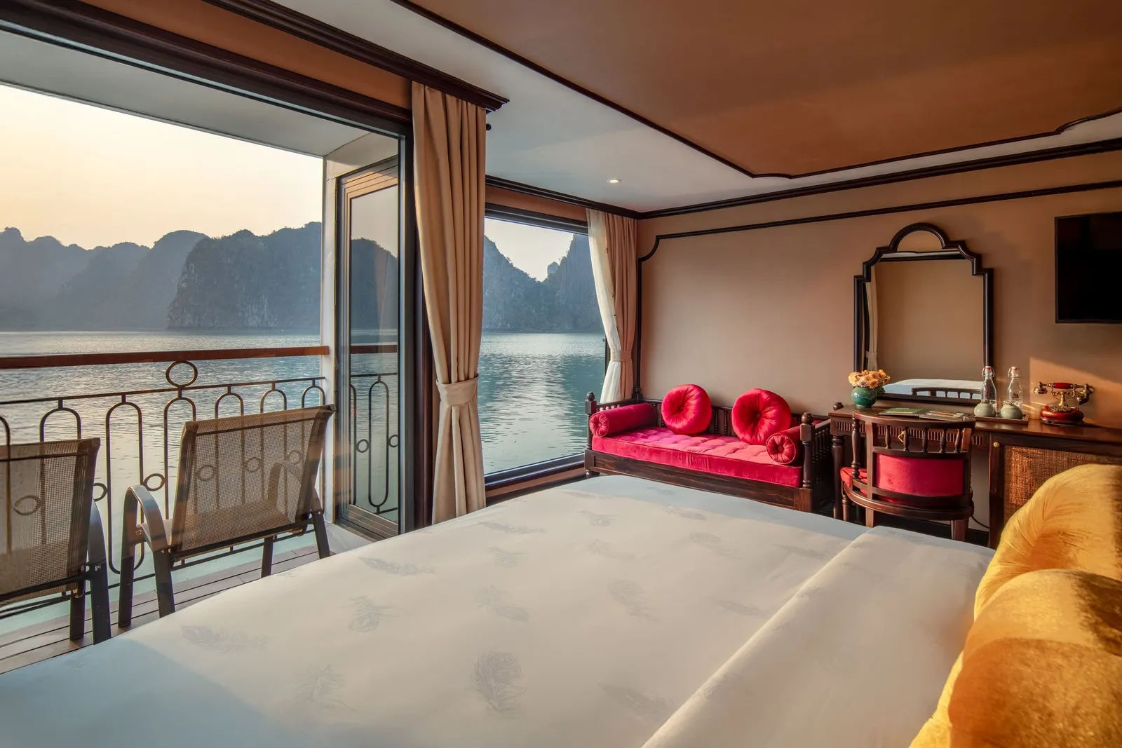 A boat cabin interior with a bed, red cushioned seating, and open balcony doors showing limestone karsts and water outside.