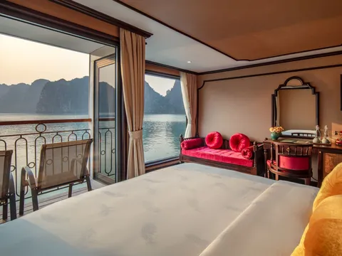 A boat cabin interior with a bed, red cushioned seating, and open balcony doors showing limestone karsts and water outside.
