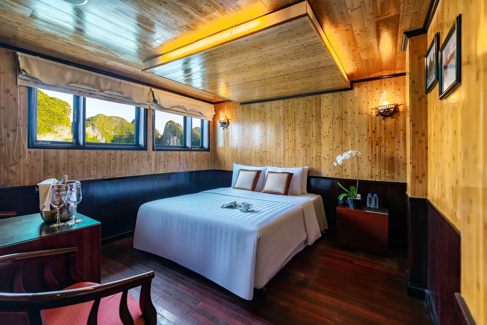A wood-paneled cabin with a white-draped double bed, wine glasses in an ice bucket, an orchid on a bedside table, and windows showing green limestone karst peaks.