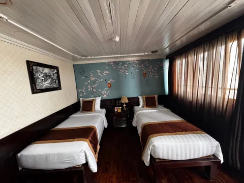 A twin cabin interior with two beds, decorative wall art, wooden ceiling, curtained windows, and a nightstand between the beds.
