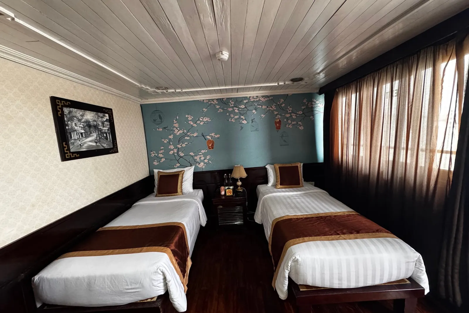 A twin cabin interior with two beds, decorative wall art, wooden ceiling, curtained windows, and a nightstand between the beds.