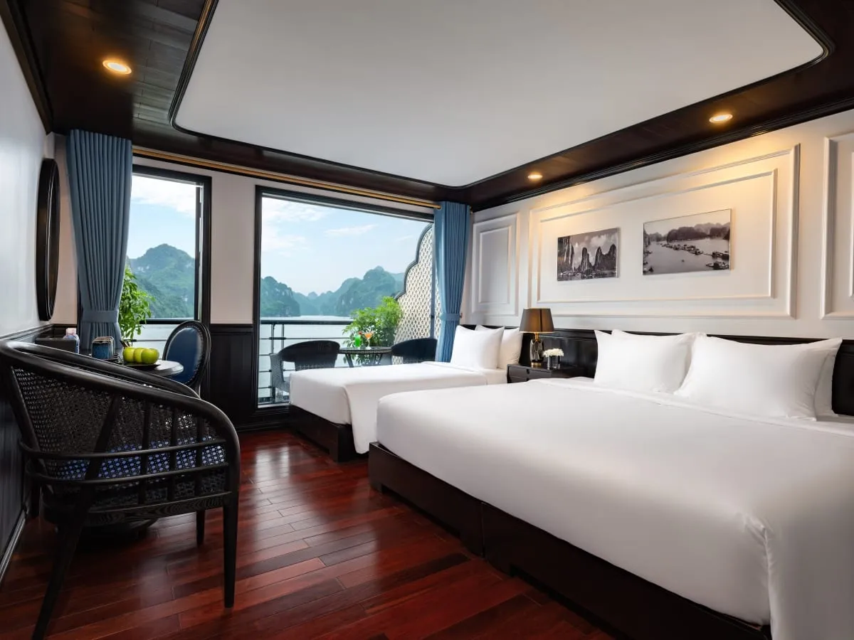A cabin with two large white beds, dark wood ceiling trim and polished wood floor, with windows and a balcony looking out on green limestone karst islands and water, and black-and-white bay photos on the wall.
