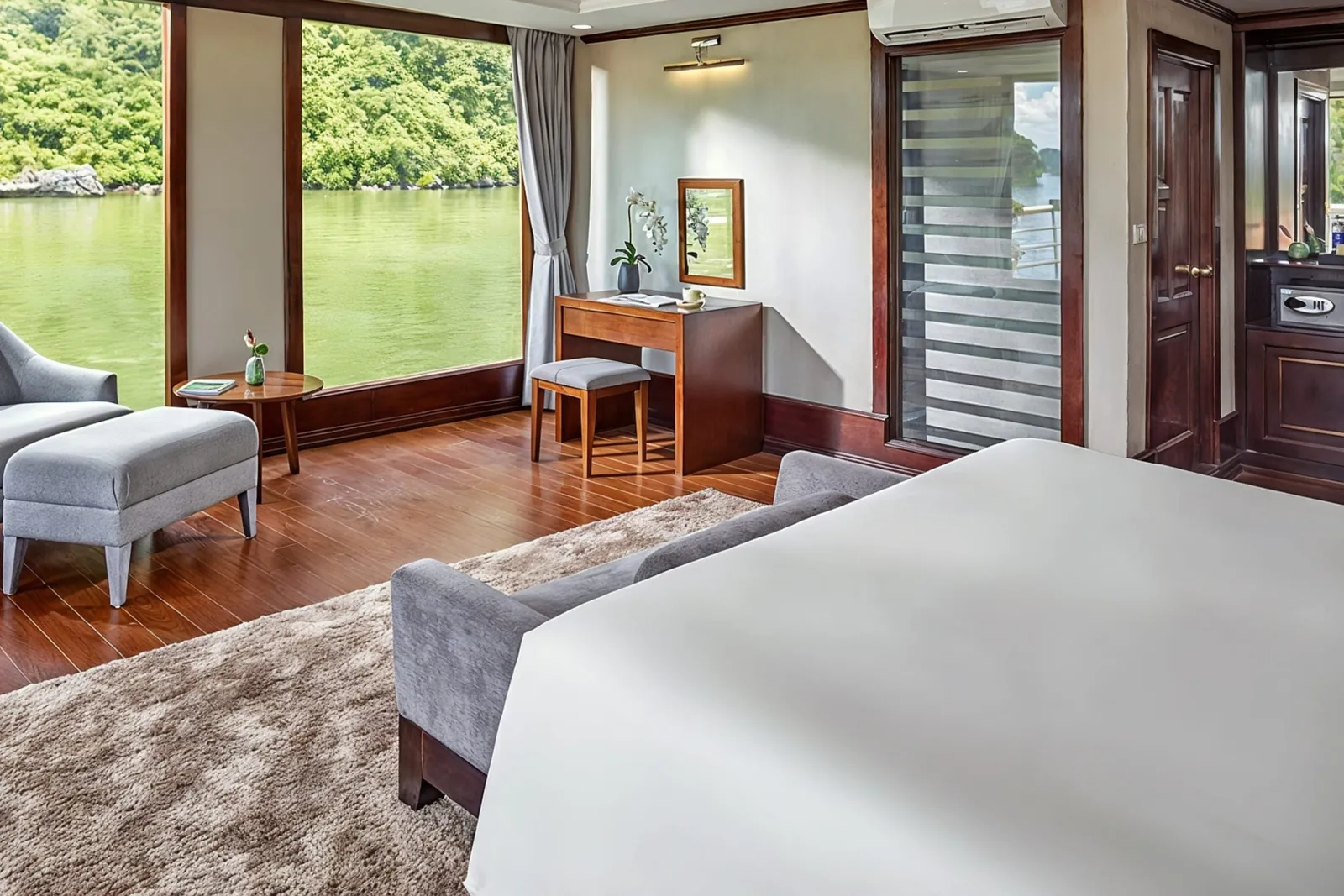 A cabin with a white bed, a grey bench, a desk and large windows facing the green bay.