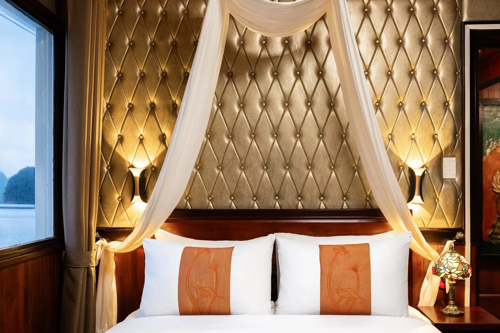 A cabin bed with white pillows bearing orange embroidered panels, a sheer white canopy, a tufted gold padded headboard wall with wall lamps, a stained-glass bedside lamp, and a window showing calm water and karst-like rock silhouettes.
