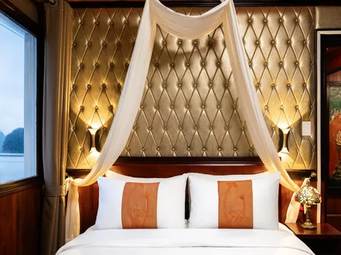 A cabin bed with white pillows bearing orange embroidered panels, a sheer white canopy, a tufted gold padded headboard wall with wall lamps, a stained-glass bedside lamp, and a window showing calm water and karst-like rock silhouettes.