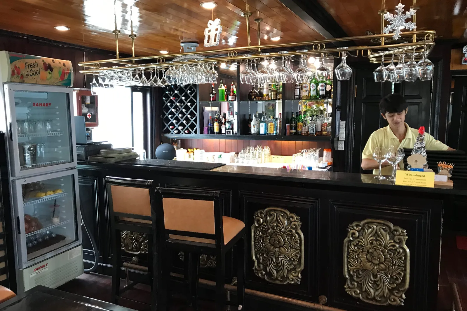 Interior bar area with ornate black cabinetry, hanging glassware, staff member in yellow shirt, and refrigerator with SANAKY branding visible.