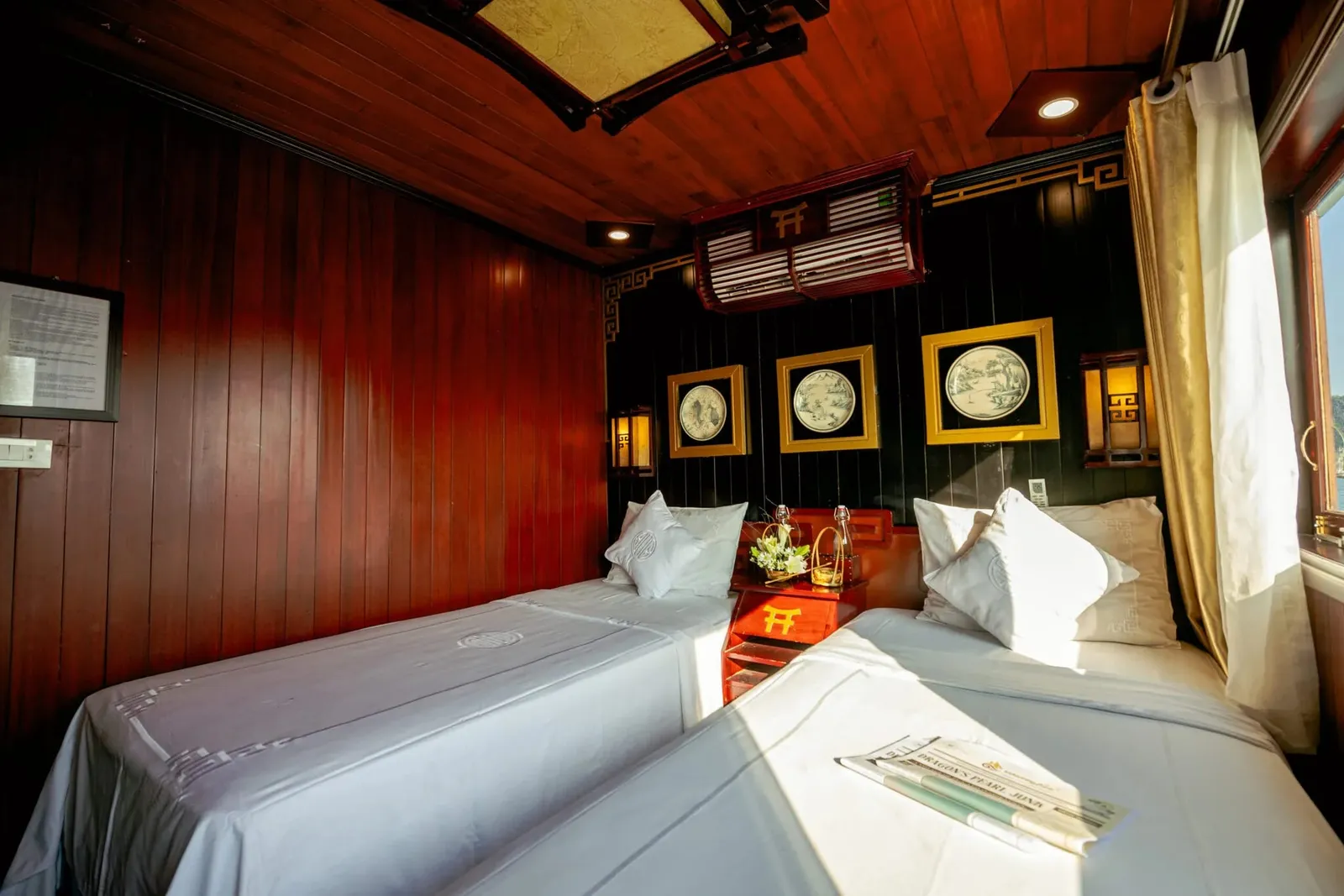 A wood-paneled twin-bed cabin with white linens, framed plates on a dark wall, red bedside table, and a brochure reading 'Dragon's Pearl Junk' on one bed, with a window showing bright outdoor light at right.