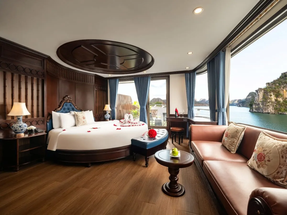 A wood-paneled cruise cabin with a round bed decorated with towel swans and rose petals, a leather sofa, and large windows overlooking green water and limestone karst cliffs.
