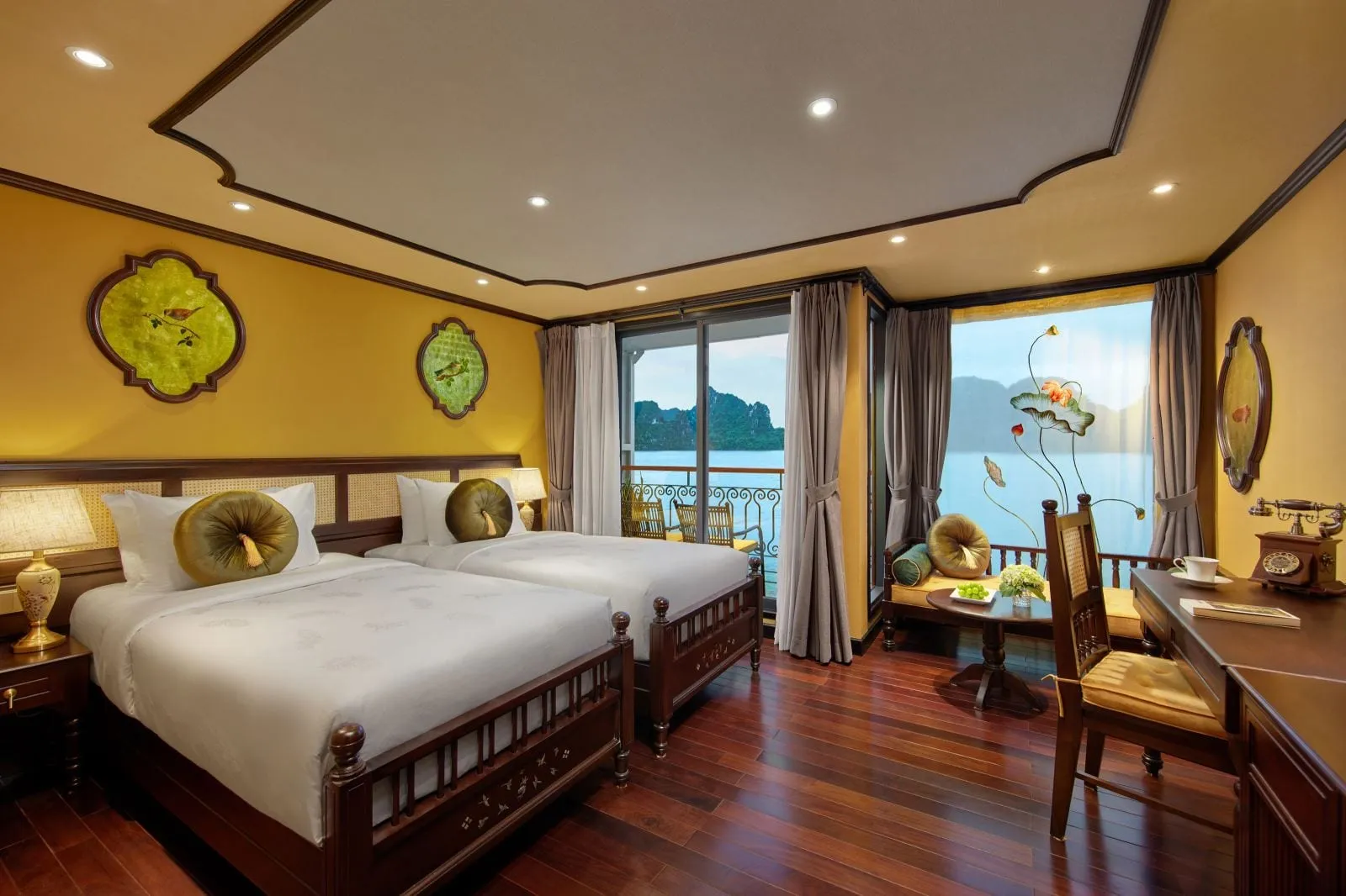 A cabin with two twin beds, yellow walls, dark wood floors and furniture, and a balcony and window overlooking calm water and limestone karst islands.