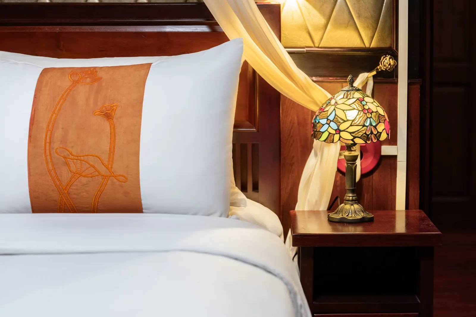 A close-up of a cabin bed with a white pillow and an orange embroidered lotus band, next to a dark wood nightstand holding a stained-glass Tiffany-style lamp, with a sheer canopy curtain and tufted padded wall behind.