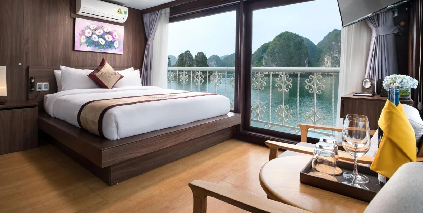A wood-paneled cruise cabin with a double bed, branded embroidered cushion and runner, and a seating area with wine glasses, with large windows and a balcony looking out on limestone karsts rising from green water.
