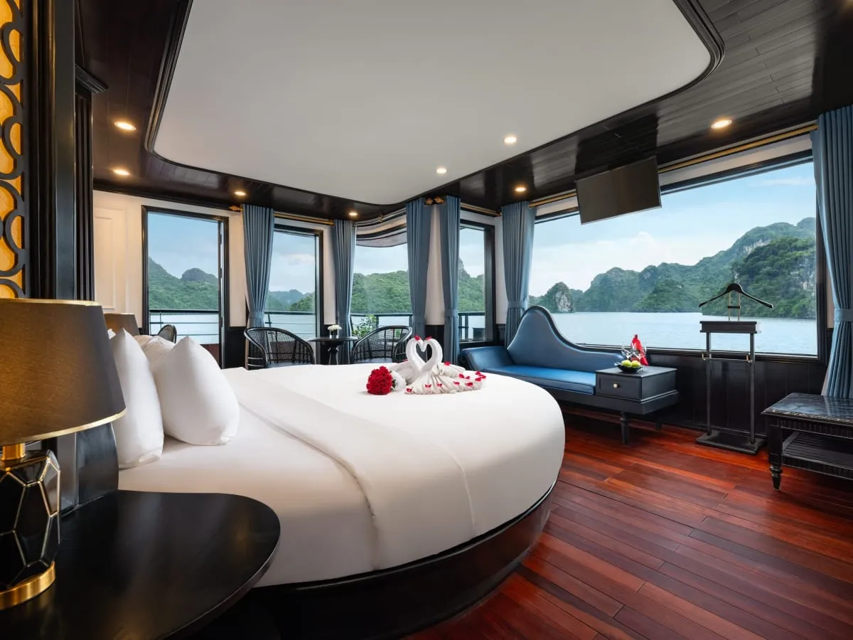 A cruise cabin with a round white bed decorated with towel swans and rose petals, a blue chaise, dark wood ceiling and red-brown wood floor, and large windows showing calm water and limestone karst islands.