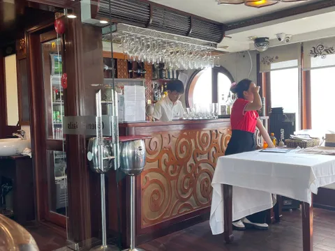 Interior bar and dining area with staff, wooden floors, decorative ceiling light, and windows showing bright exterior light.