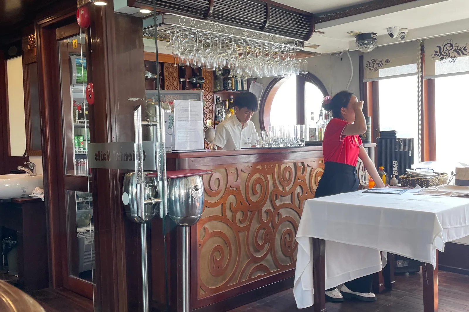 Interior bar and dining area with staff, wooden floors, decorative ceiling light, and windows showing bright exterior light.