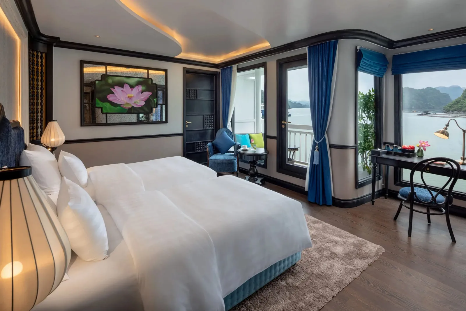 A cruise cabin with a large white bed, blue curtains, a lotus artwork on the wall and a desk, with windows and a balcony door opening onto a bay of calm water and forested limestone hills.