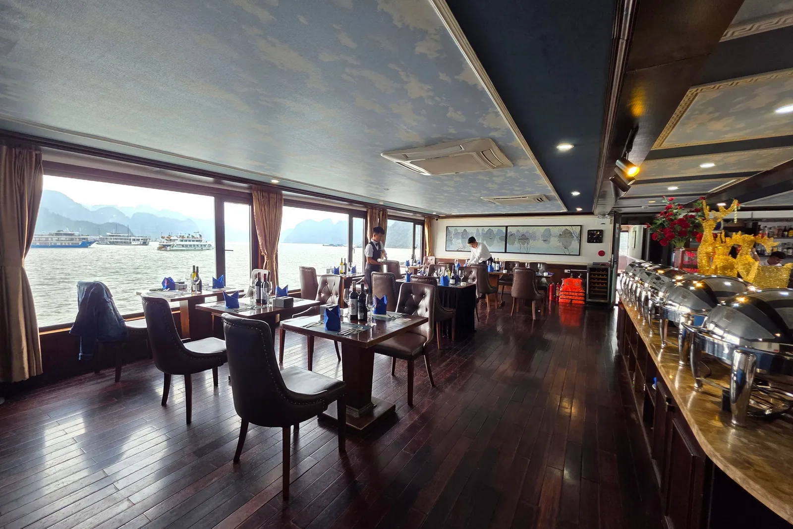 Interior dining room of a cruise boat with set tables, buffet area, and windows showing karst limestone formations and other boats on the water.