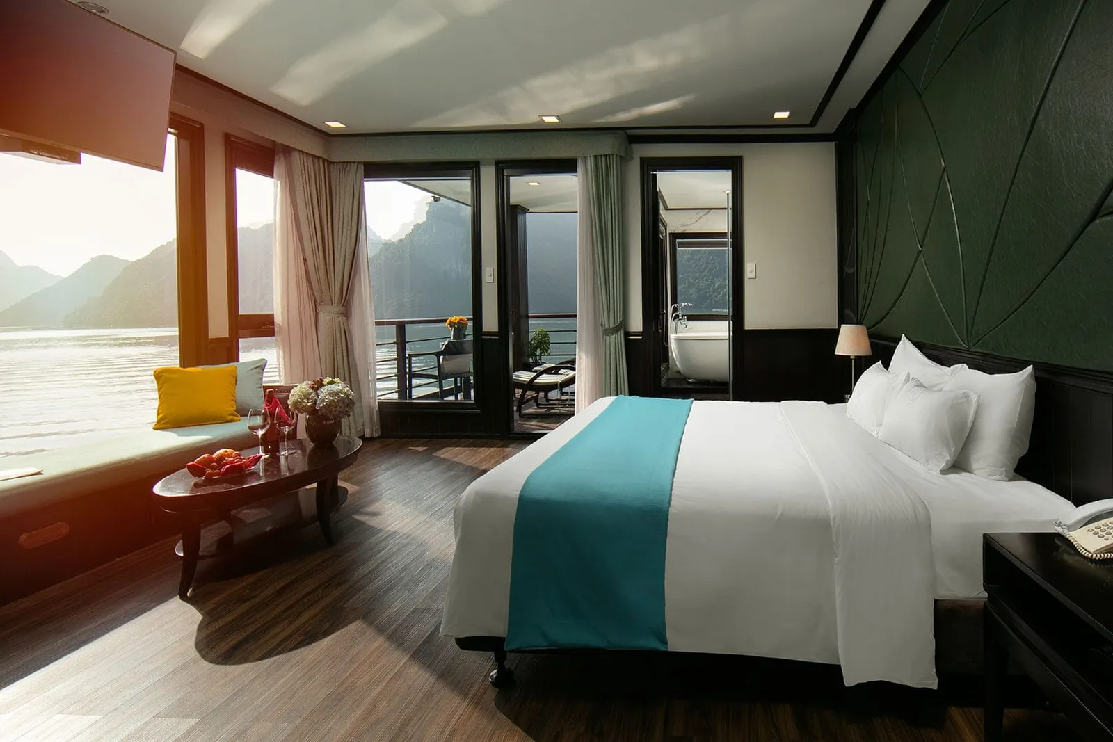 A luxury boat cabin with a bed, seating area, and large windows showing karst mountains and calm water outside.