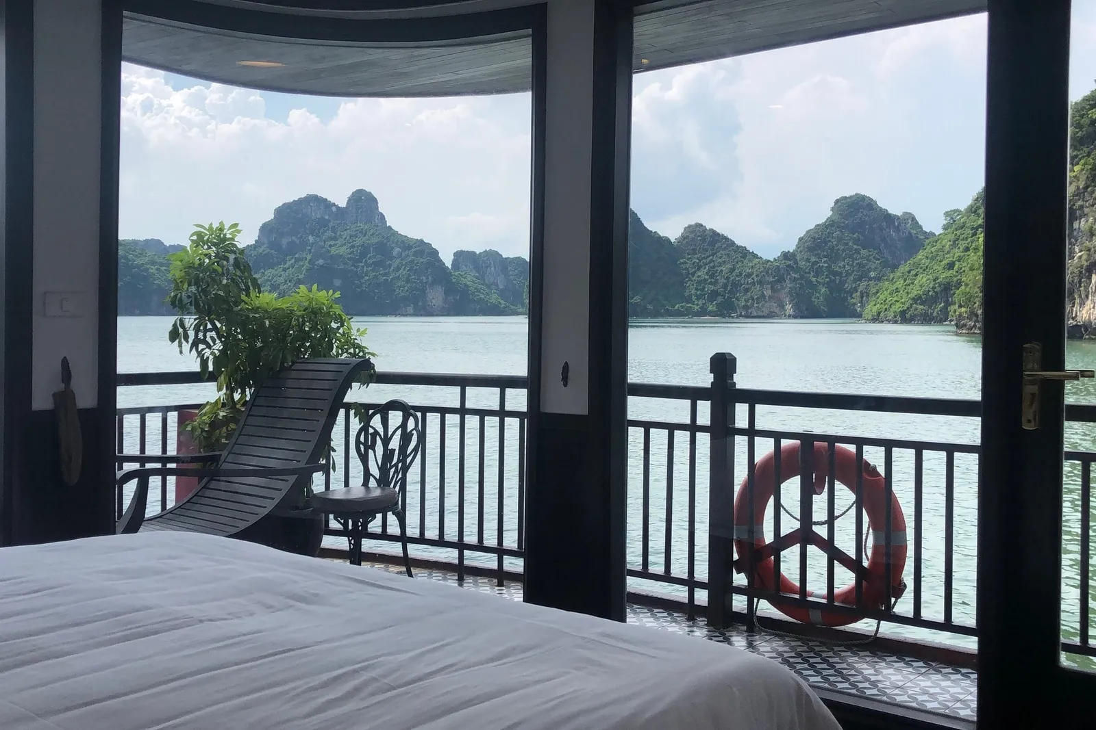A boat cabin interior with a bed, lounge chair, and balcony railing overlooking limestone karsts and green water.