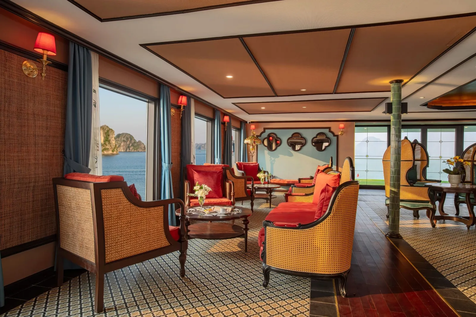 An elegant boat interior lounge with rattan chairs, red cushions, and windows showing limestone karsts and water outside.