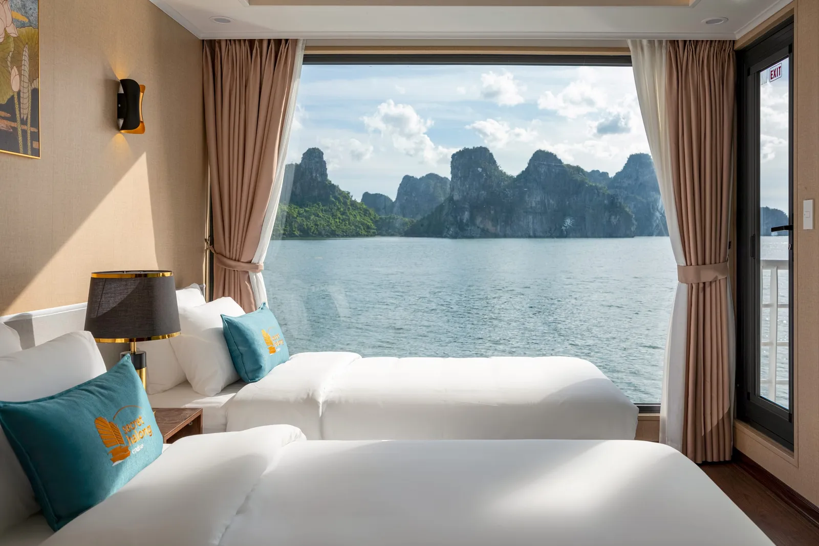 Twin beds with white linen and teal cushions beside a large window with curtains, with limestone islands outside.