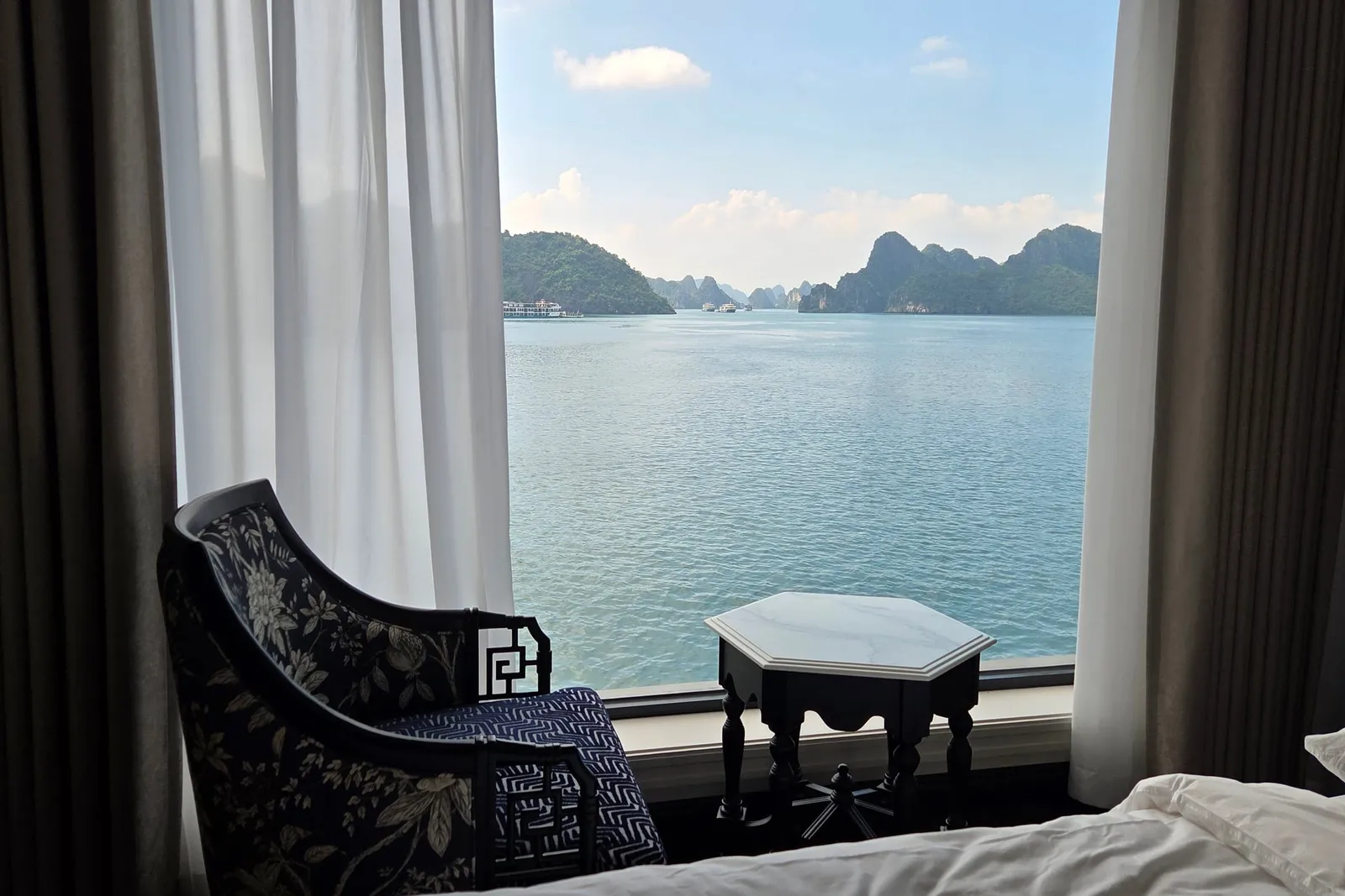 A cabin interior with a patterned armchair, a small hexagonal side table, curtains and the edge of a bed in front of a large window overlooking calm water and forested limestone karst islands.