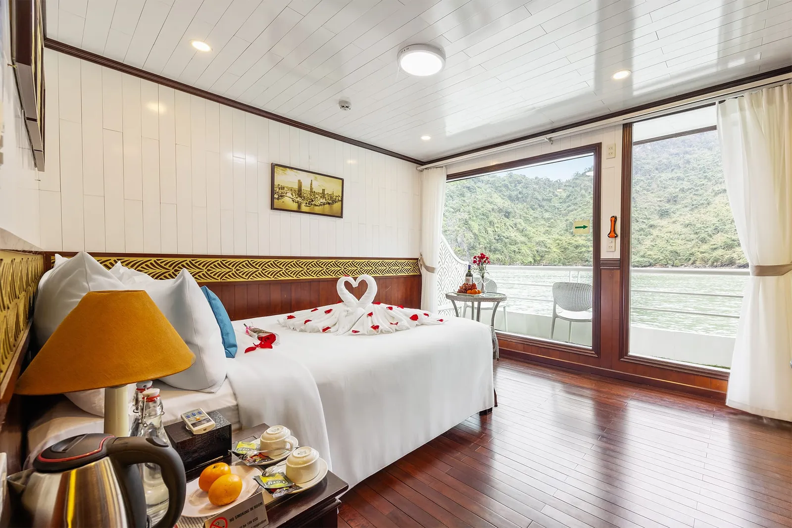A cruise cabin with a double bed decorated with a towel-swan heart and rose petals, wooden floor, and glass doors opening to a balcony overlooking calm water and forested limestone hills.