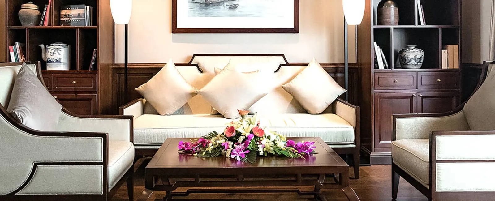 A wood-paneled lounge with a cream sofa, two matching armchairs, cushions, a low coffee table with a flower arrangement, bookshelves with ceramics, and a framed picture on the wall.