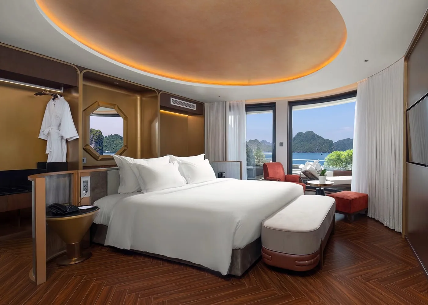 A luxurious cruise cabin with a king bed, herringbone wood floor, gold-toned walls, a circular lit ceiling, and large windows overlooking karst limestone islands on the water.