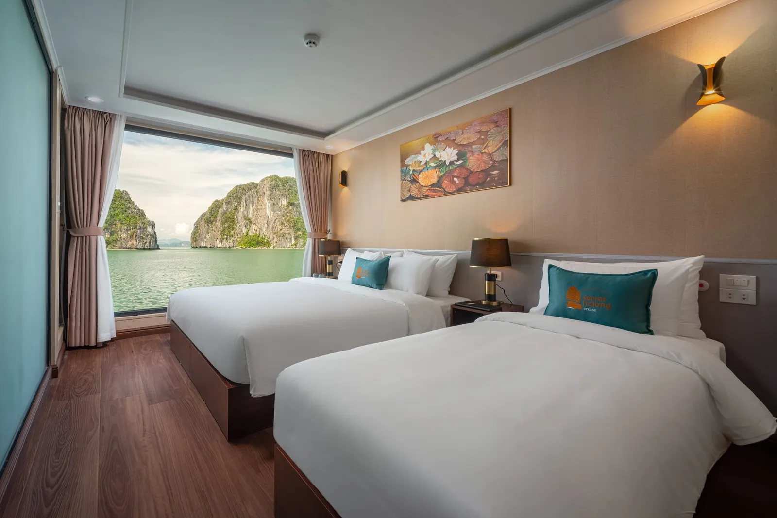 A cabin with a large window facing the bay between brown curtains, with white beds, a wall lamp and a framed painting.