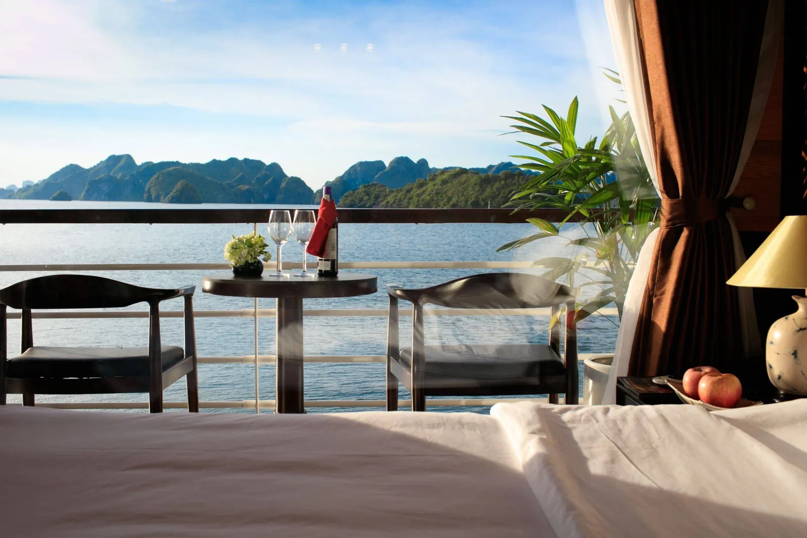 View from a bed in a cabin through open balcony doors to two chairs and a small table with wine and glasses, a railing, calm water and green karst islands beyond.