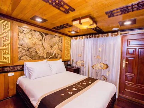 A cruise boat cabin interior with a double bed, carved wood wall art depicting cranes, decorative wooden ceiling panels with recessed lighting, embroidered curtains with lotus motifs, and a dark wood door.