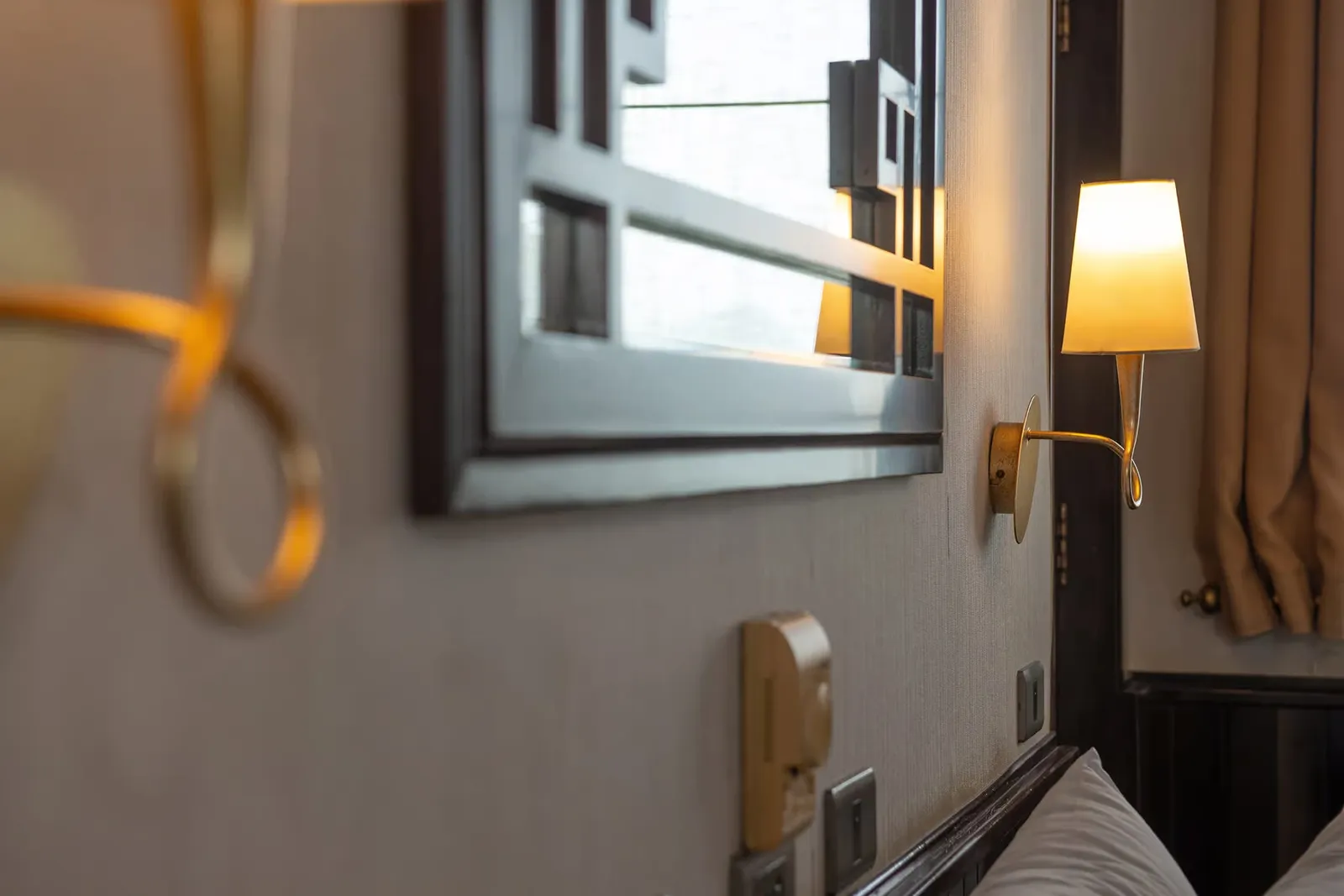 Close-up of a cabin wall with a framed mirror reflecting a lit lamp, a gold wall sconce with a cream shade, wall switches, a pillow edge, and beige curtains.