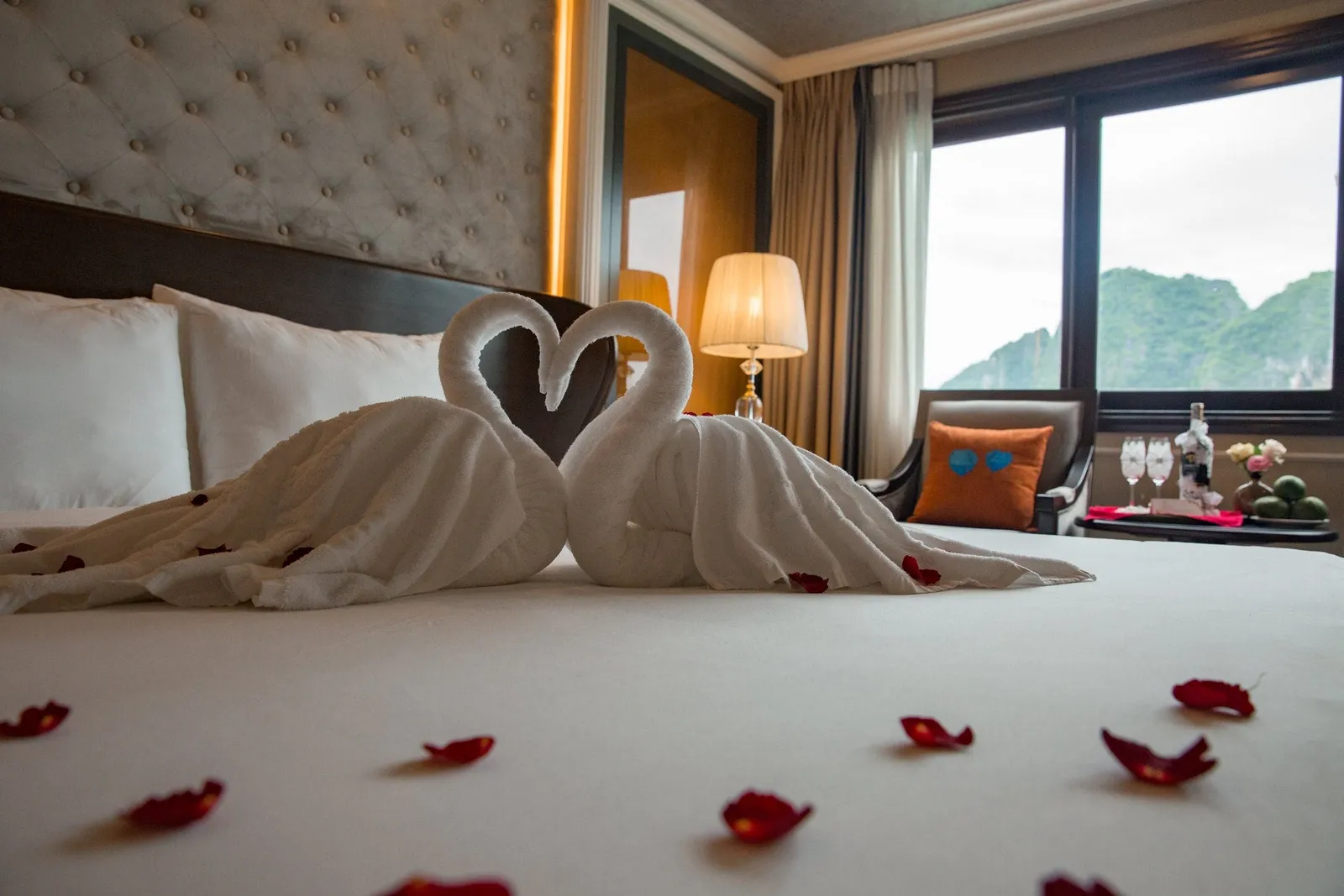 A cabin bed with two towel swans forming a heart and scattered red rose petals, with a window showing green limestone karst hills.