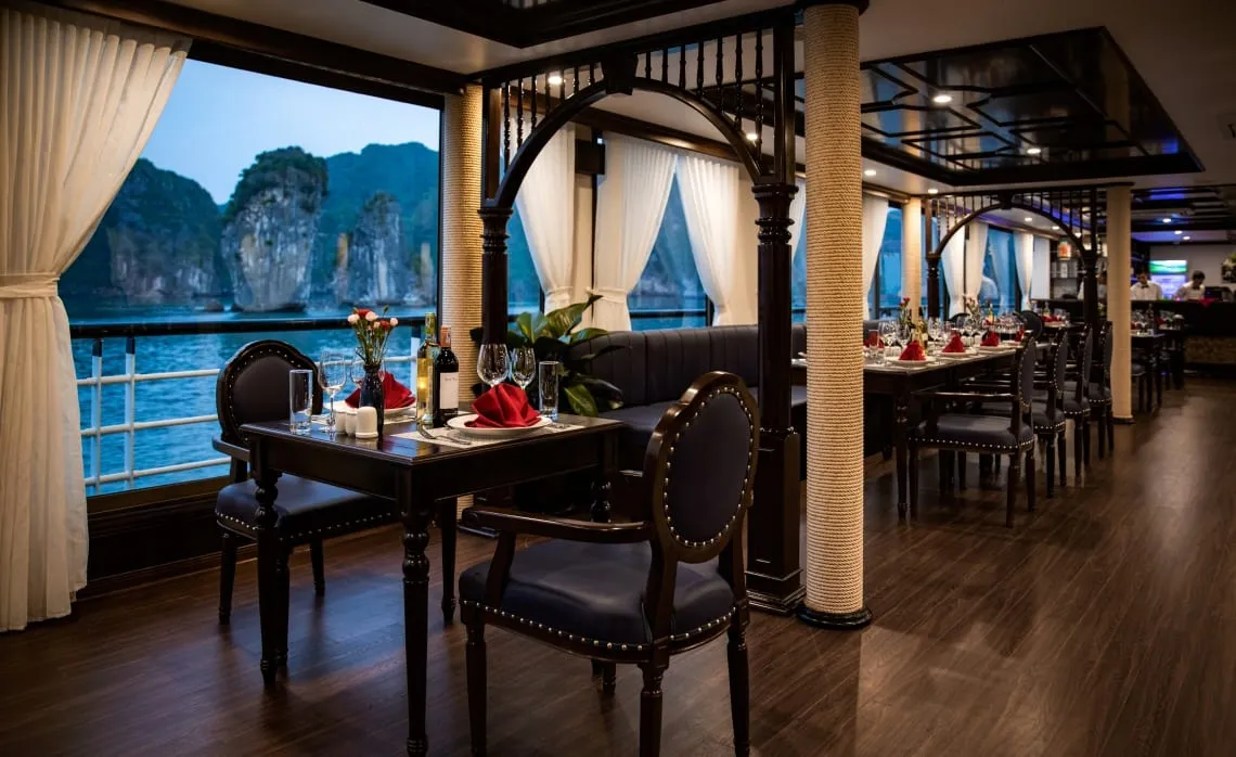 Interior of a boat dining room with dark wood tables set with red napkins and wine glasses, rope-wrapped pillars, white curtains, and a window view of limestone karsts on calm water at dusk.