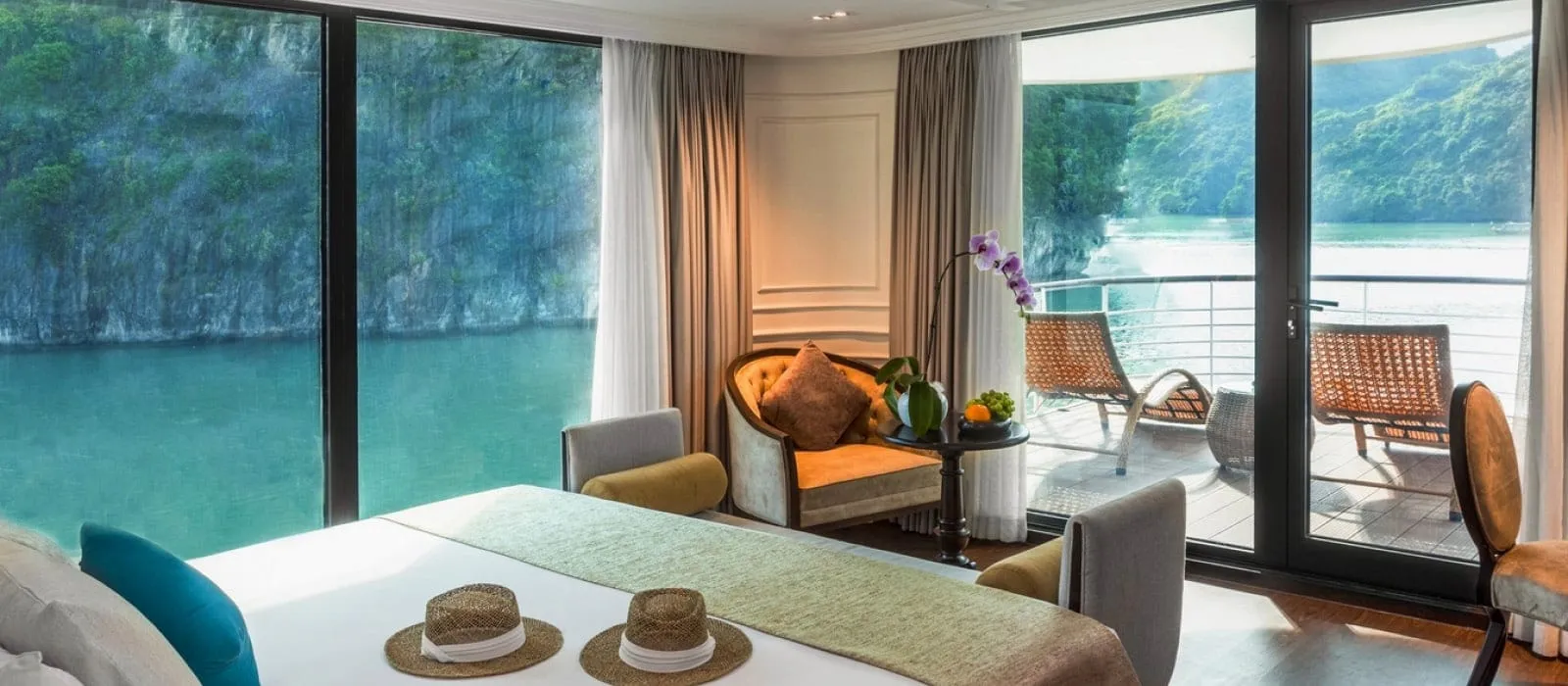 A cruise cabin interior with a bed, armchair, orchid and a balcony with rattan chairs, with floor-to-ceiling windows looking out on green water and limestone karst cliffs.