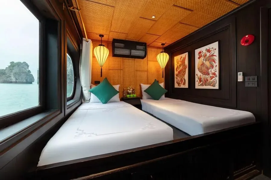 A twin-bed cabin with white linens, teal cushions, bamboo-weave walls and ceiling, silk lanterns and two folk-art prints, with a window showing calm green water and a limestone karst island.