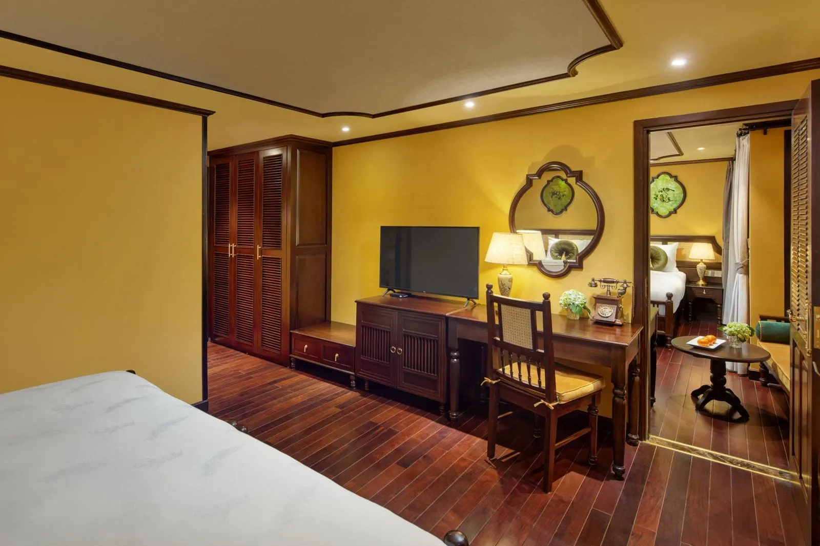 A yellow-walled wood-paneled cabin with a bed in the foreground, a desk with TV, vintage telephone and chair, and a doorway opening to a second room with another bed and a small round table.