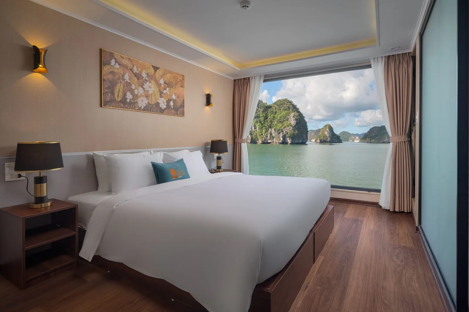 A cabin with a double bed in white linen and a teal cushion, a tray ceiling with warm lighting, a framed painting and a bay-view window.