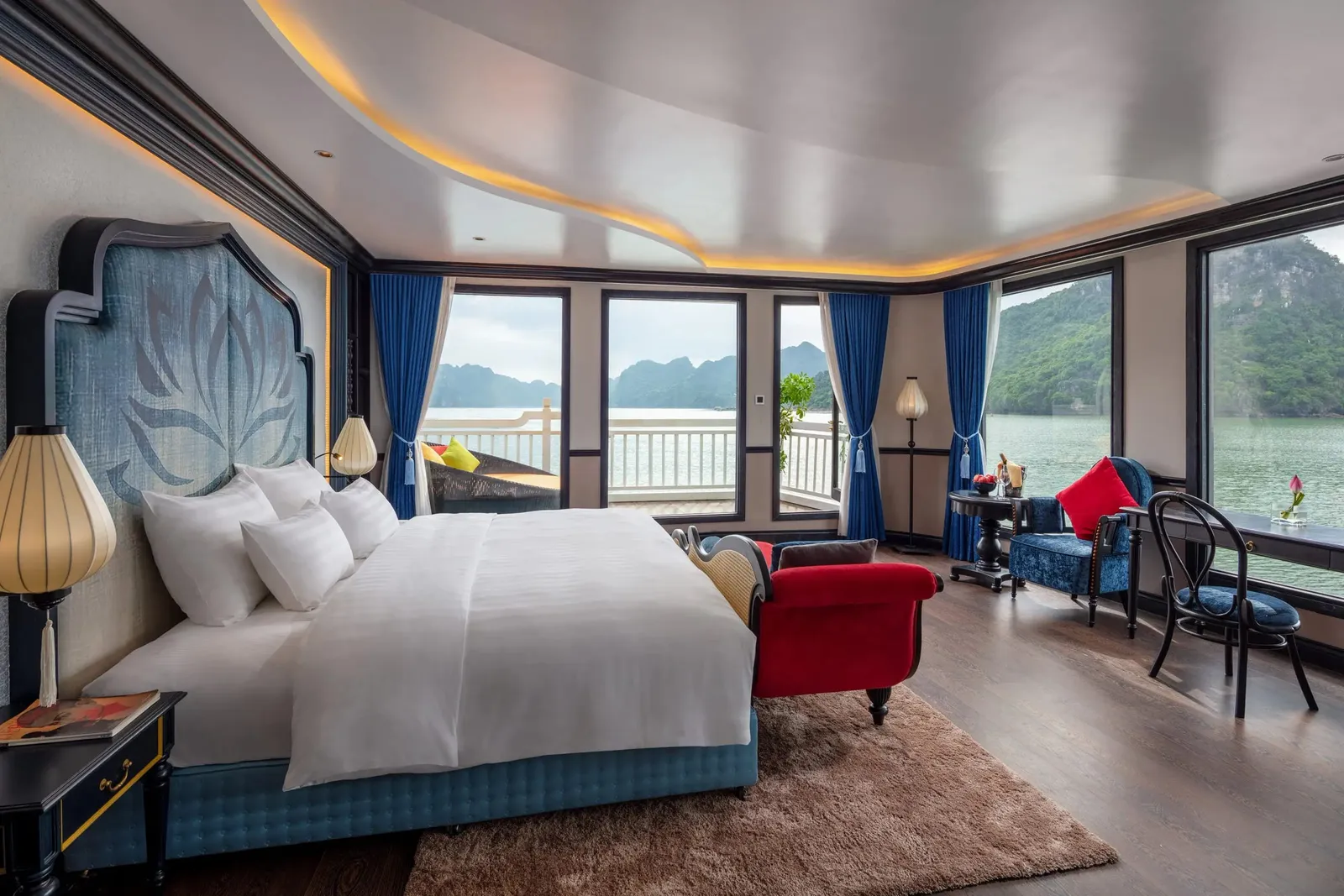 A cruise cabin with a large white-covered bed, lotus-motif headboard, red bench, blue armchair and desk, with floor-to-ceiling windows and a balcony overlooking green water and limestone karst islands.