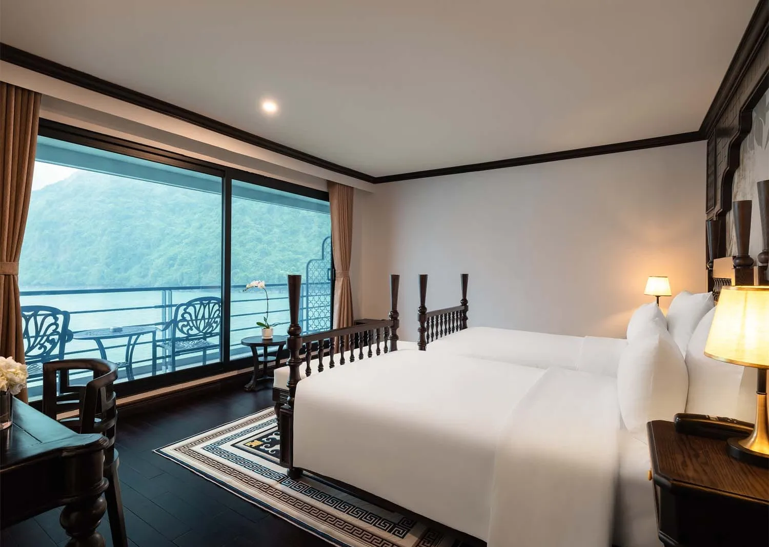 A twin-bed cruise cabin with dark wood furniture, a patterned rug, warm lamp lighting, and a balcony overlooking green karst hills and calm water.