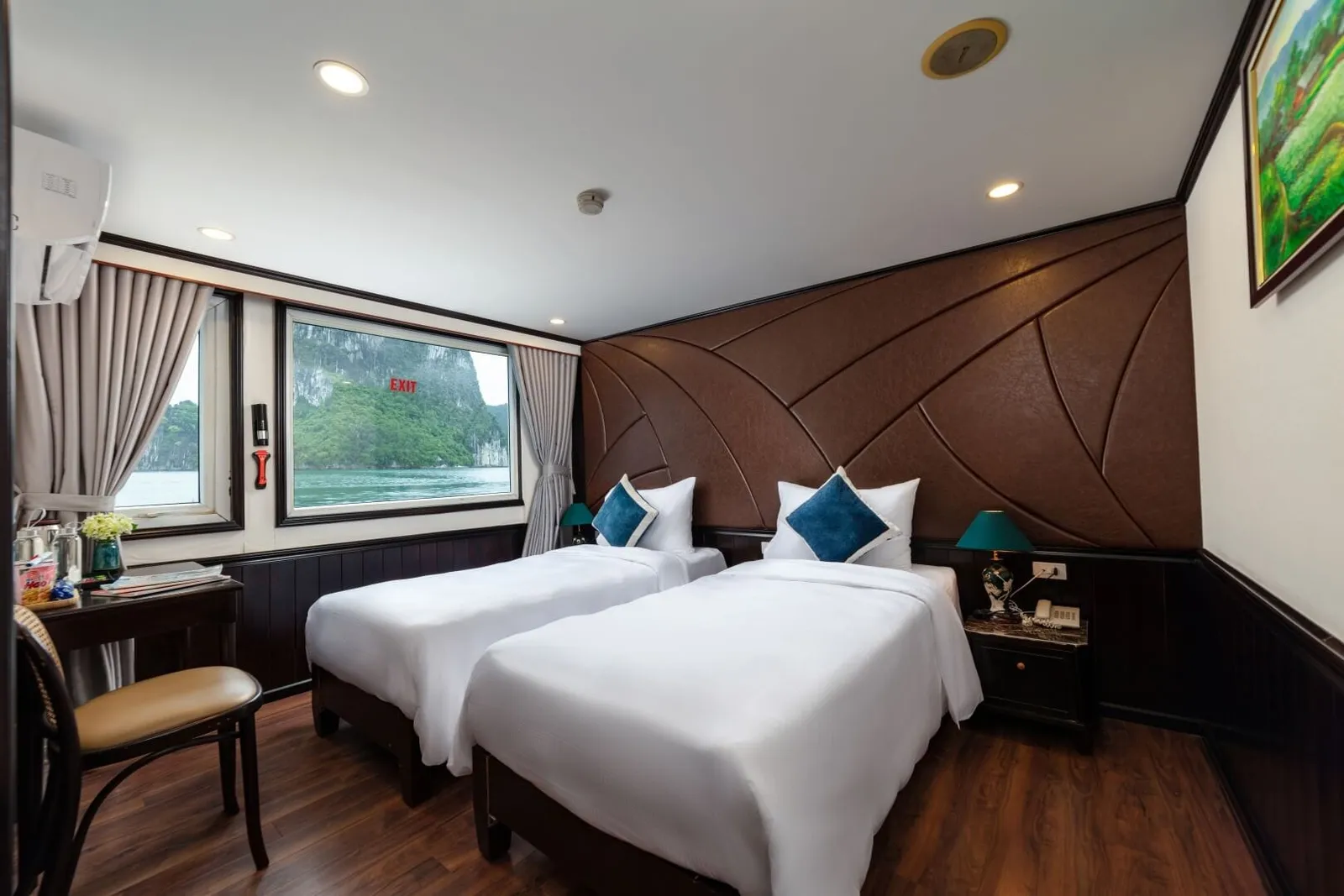 A cabin with two twin beds in white linen with blue cushions, a brown leather-paneled headboard wall, wooden floor, and a window showing limestone karst cliffs and calm green water.