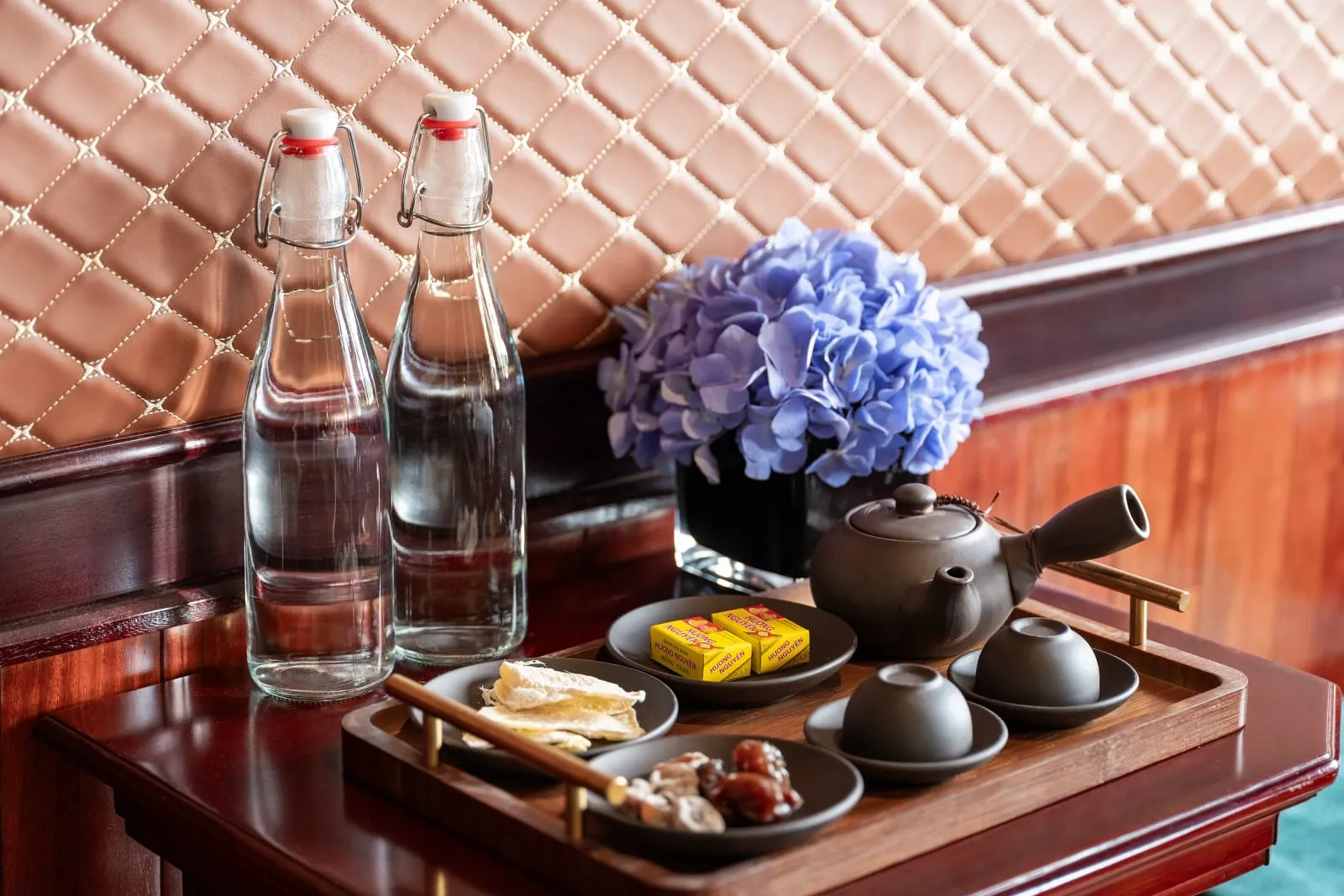 A wooden tray with a dark clay teapot, two cups, small dishes of dried ginger and dried fruit, and wrapped sweets sits on a polished wood table beside two glass water bottles and a blue hydrangea arrangement, in front of a quilted tan leather wall panel.
