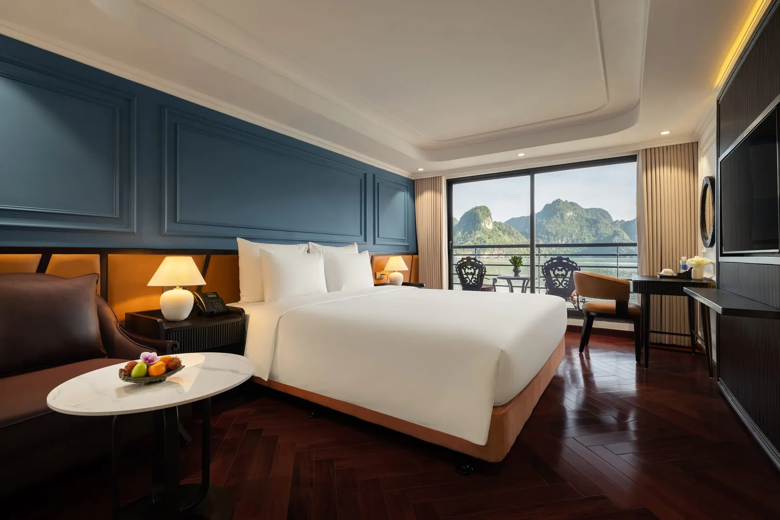 A luxury cabin interior with a large bed, dark wood flooring, and floor-to-ceiling windows showing limestone karst islands and water outside.