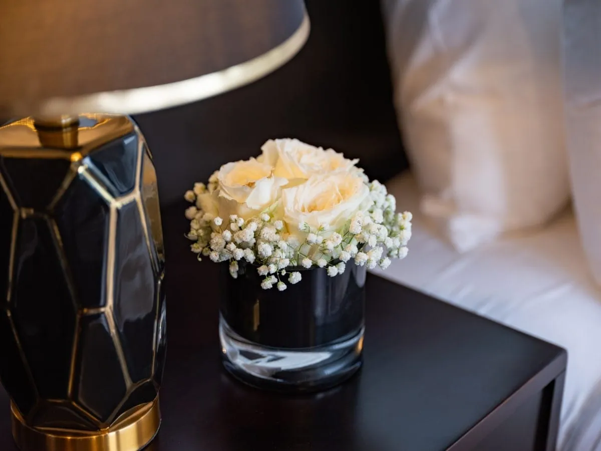 A small black vase of cream roses and baby's breath sits on a dark bedside table next to a black and gold faceted lamp, with white pillows and bedding behind.